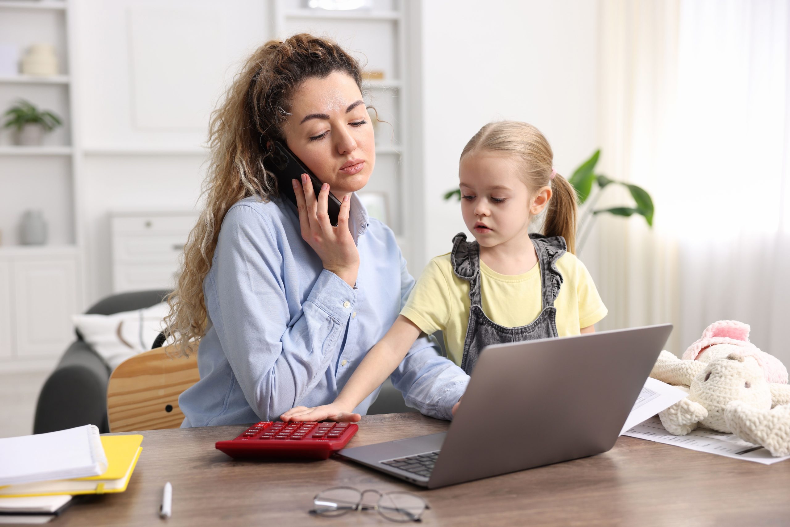 Vrouw bekijkt haar financiën met een laptop en rekenmachine terwijl haar dochter naast haar zit